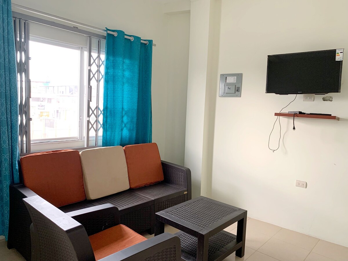 A small living area is presented, featuring a dark wicker sofa with orange and cream cushions. A square coffee table sits in front, while a wall-mounted television is positioned on a shelf. Bright blue curtains frame the window, allowing natural light to illuminate the space.