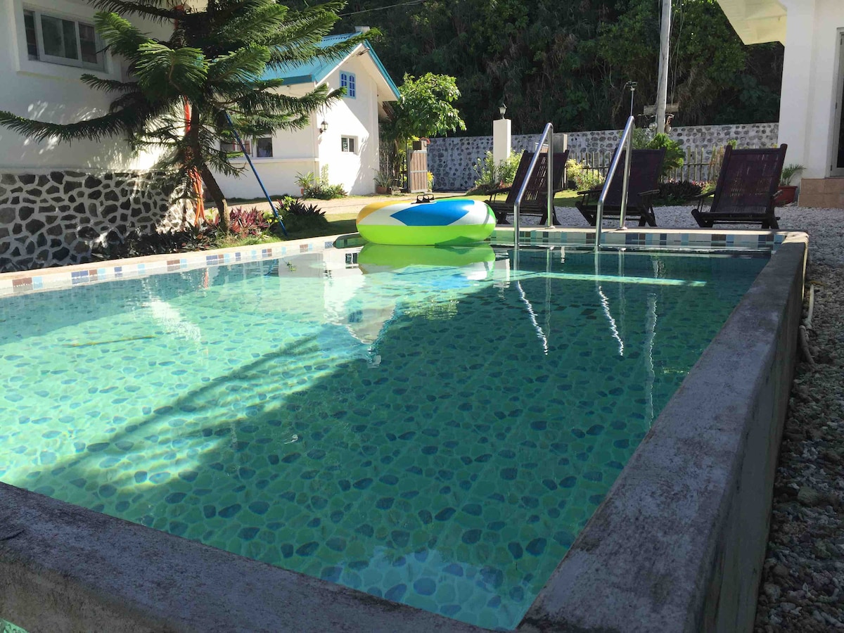 A refreshing pool is framed by a vibrant green environment, featuring a colorful float resting on the water's surface. Comfortable chairs are positioned alongside the pool, inviting relaxation beneath natural shade.