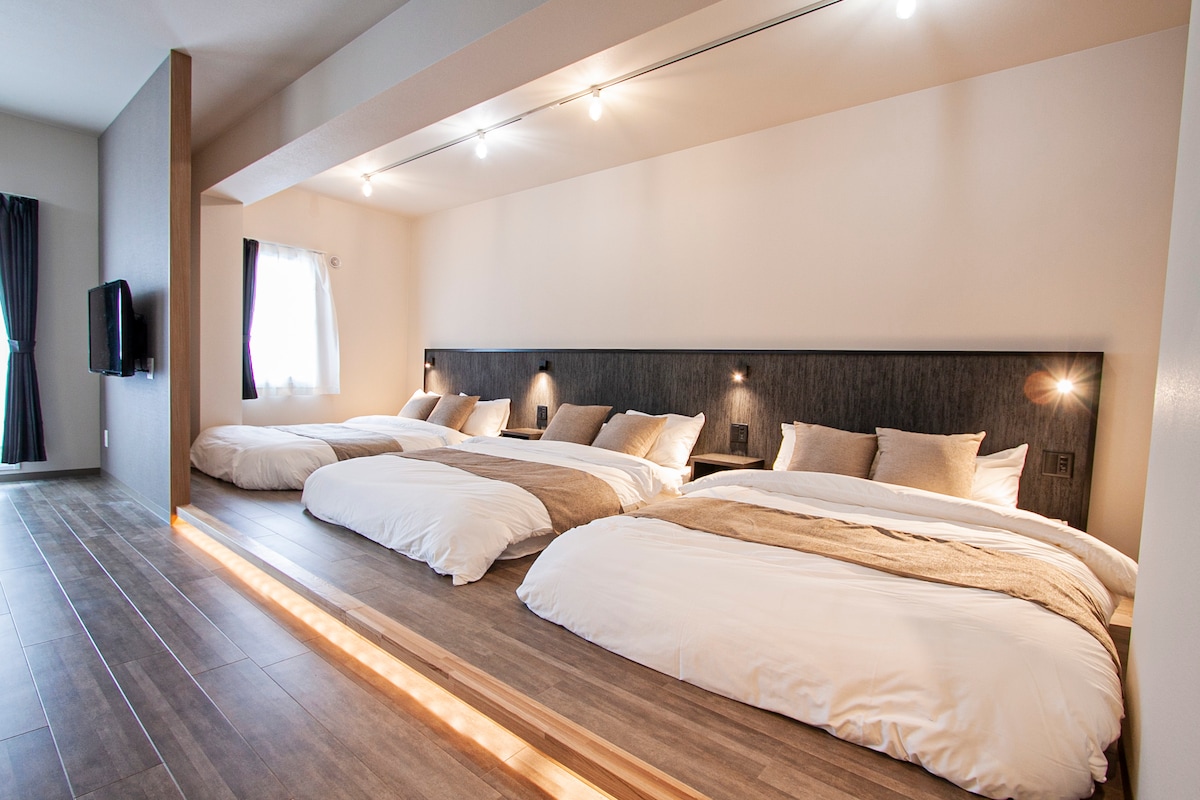 A spacious sleeping area is equipped with three large beds, dressed in soft white linens. Warm-toned lighting highlights the minimalist decor. A window offers natural light, partially covered by dark curtains, and a television is mounted on the wall.