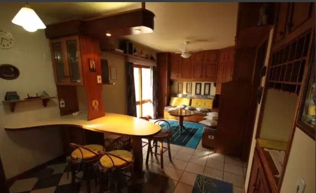 A spacious living area is furnished with wooden cabinetry and a cozy seating arrangement. A light-colored bar table with seating is visible, alongside large windows that allow natural light to illuminate the room.