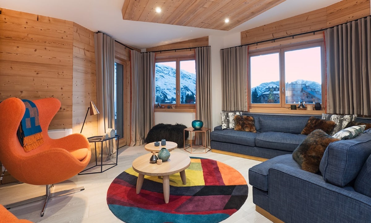 A spacious living area features a blend of cozy seating, including an orange armchair and a dark sectional sofa. Large windows provide views of the snowy landscape outside, while a vibrant area rug adds color to the neutral-toned decor. Soft lighting enhances the inviting ambiance.