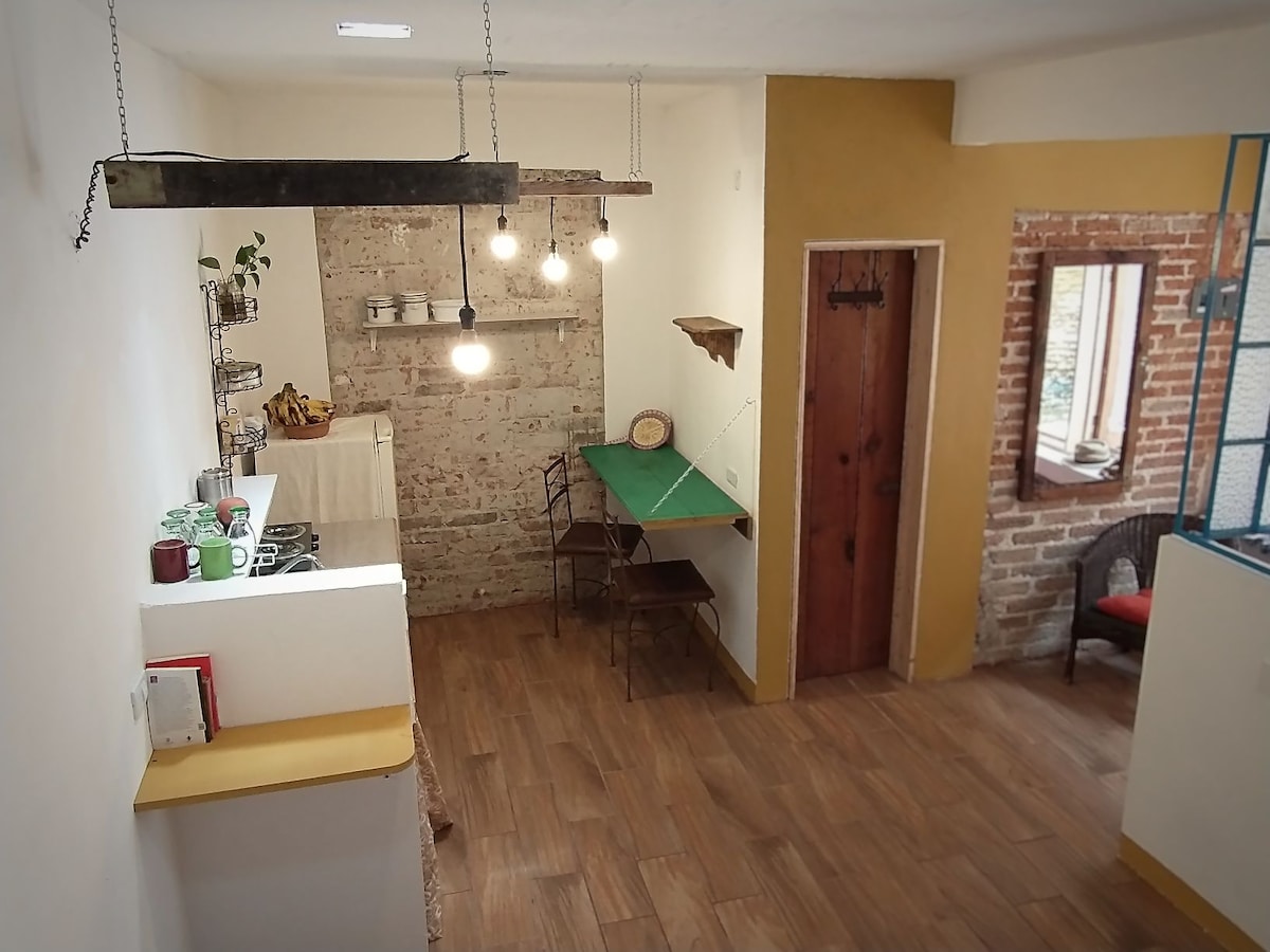 A rustic kitchen workspace features exposed brick walls complemented by warm yellow accents. Industrial-style lights hang from the ceiling above a small green table with two chairs. A wooden door and a nearby window introduce natural light, creating a cozy atmosphere.