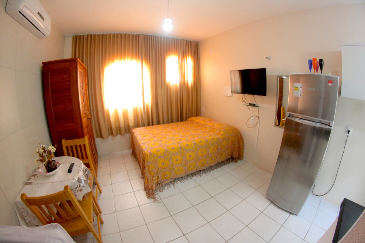 A spacious guest room features a double bed covered with a patterned blanket, complemented by a nearby wardrobe and a wall-mounted TV. A dining table with two chairs is positioned beside a window, while the kitchen area includes a refrigerator and minimal appliances.