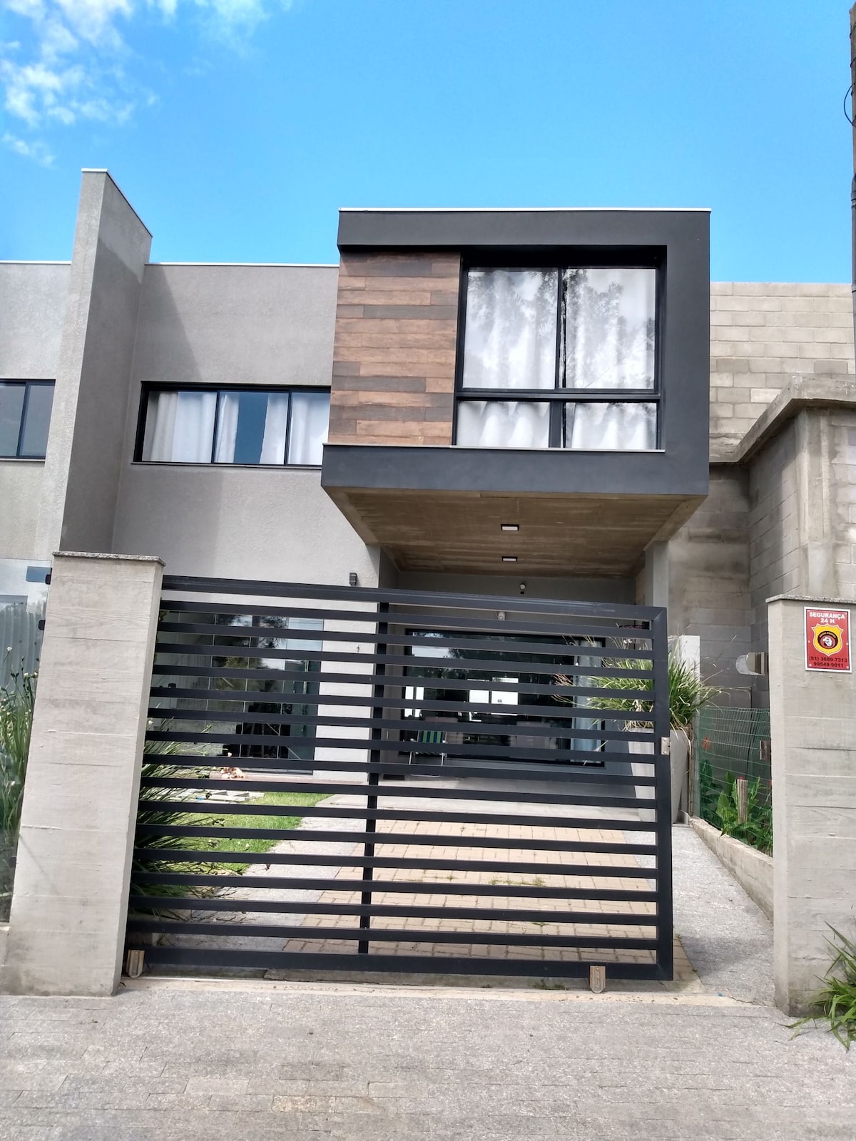 The exterior of a modern building is featured, showcasing a blend of grey and wood accents. A black metal gate offers access to the property. Large windows provide natural light, and a clear blue sky is visible above.