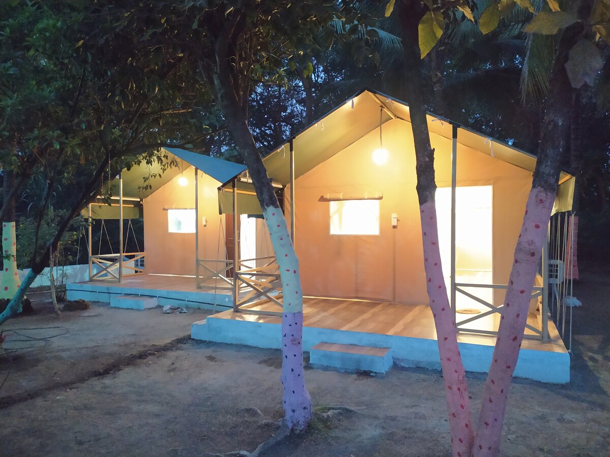 Two spacious canvas tents are nestled under trees, softly illuminated by warm lights. Each tent features large windows allowing natural light to filter in. A wooden platform supports the tents, enhancing their distinct appearance in a serene outdoor setting.