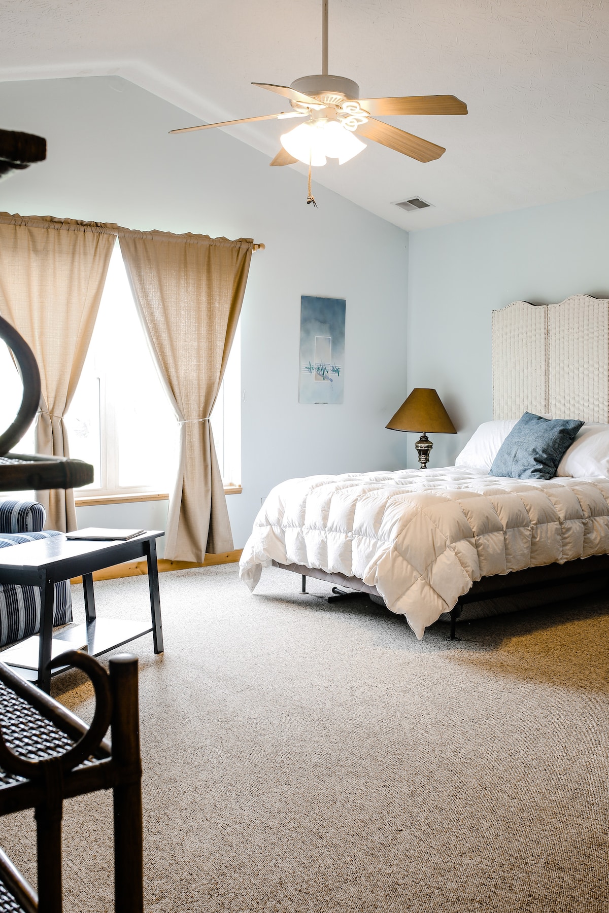A spacious bedroom features a large bed with a soft white comforter. Natural light filters through two curtain-covered windows, illuminating the room. A ceiling fan and a bedside lamp provide functional lighting. A chair and a small table are also visible in the cozy corner.