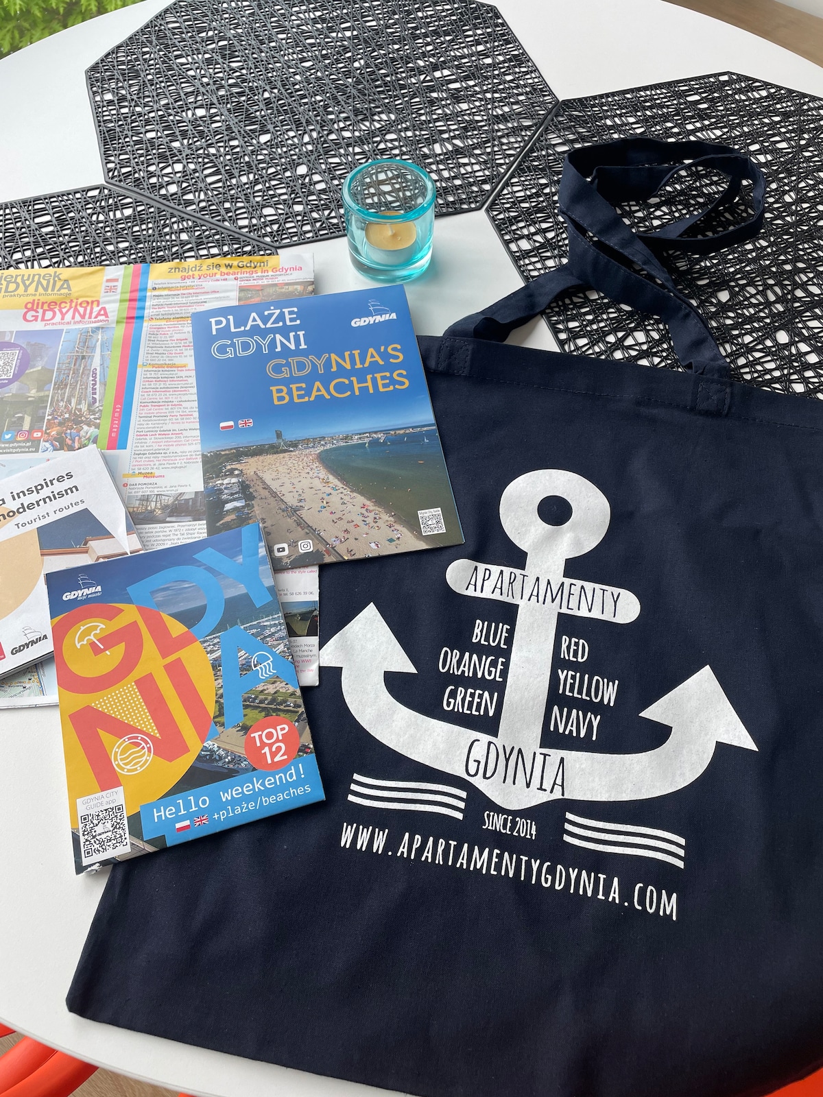A navy tote bag with a white anchor design is placed on a textured surface. Nearby, various brochures and maps about Gdynia are arranged, showcasing local attractions and beaches. A small candle sits atop a decorative coaster, adding to the scene.