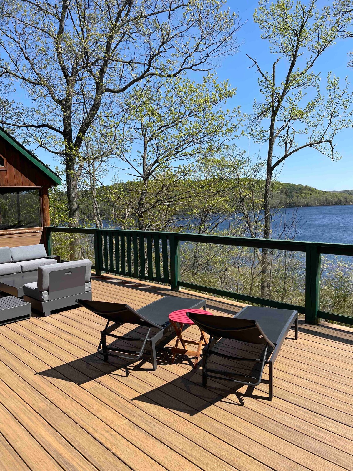 An outdoor deck is presented, featuring comfortable seating arrangements including a grey lounge set and two modern black chairs. A small red table contrasts with the wooden flooring. The backdrop reveals a tranquil lake surrounded by greenery under a clear blue sky.