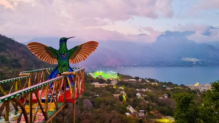 A colorful mural of a hummingbird is displayed on a viewing platform overlooking the mountainous landscape and a body of water. The surrounding hills are adorned with greenery, and distant lights reflect on the lake, creating a serene atmosphere.