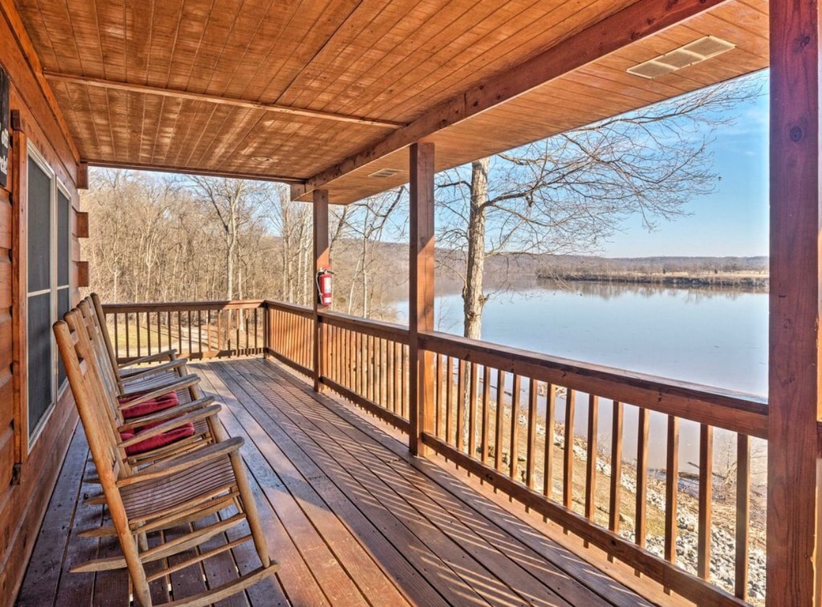 A spacious porch features several rocking chairs arranged for relaxation, with expansive views of the water in the distance. The wooden planks create a warm, inviting atmosphere, while the surrounding trees add a natural touch to the tranquil setting.