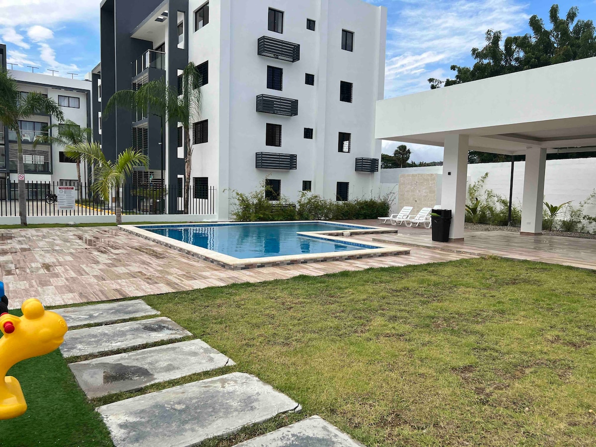 An outdoor area features a rectangular swimming pool surrounded by lounge chairs. Freshly cut grass complements the tiled decking, while palm trees add a touch of greenery. A playful structure, resembling a small animal, is situated to one side, enhancing the family-friendly environment.