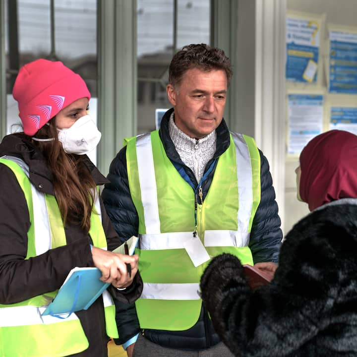 Two people in bright yellow vests listen to a person in a red head covering and fluffy black jacket.