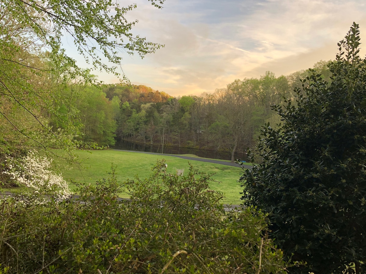 A serene landscape is visible, with lush green grass bordered by trees in full leaf. A gentle slope leads to a tranquil pond, reflecting the colors of the sky at sunset. The area is surrounded by a mixture of foliage, creating a peaceful and natural atmosphere.
