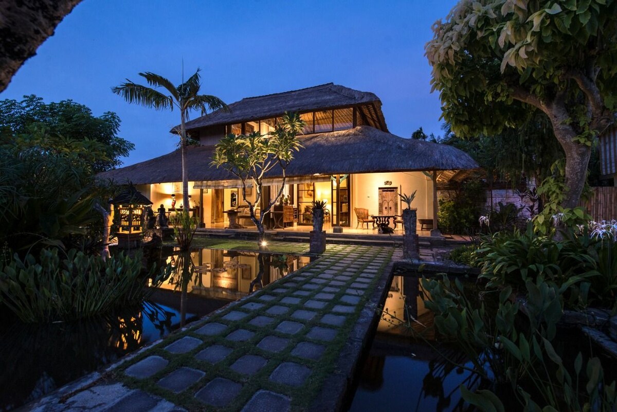 A traditional Balinese cottage is showcased against a twilight sky, with a thatched roof and expansive windows. A stone pathway leads across a reflective pond, flanked by lush greenery and tropical plants, creating a serene entryway to the inviting atmosphere of the property.