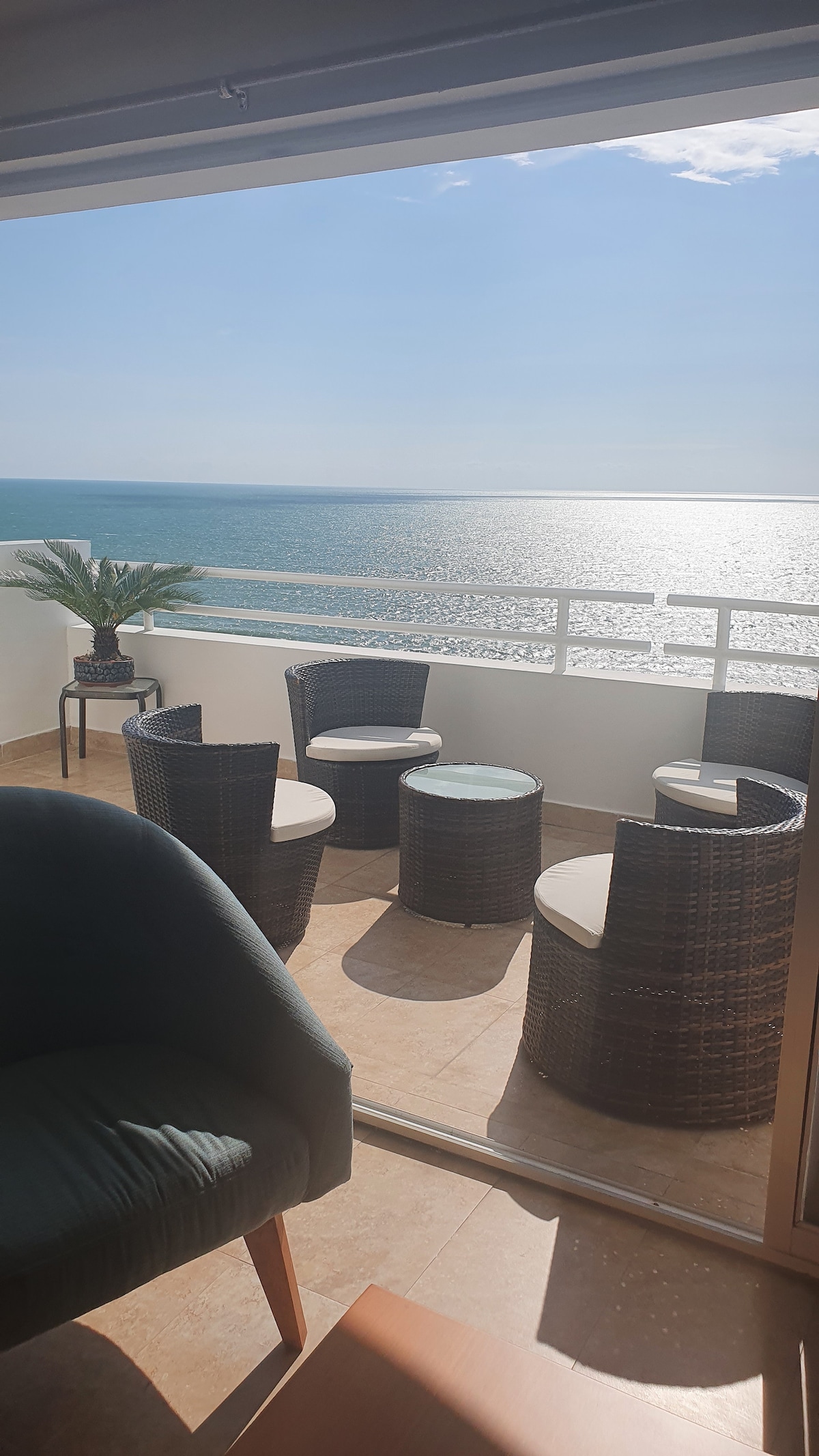 A balcony presents a serene seating area with four wicker chairs and matching tables, overlooking the ocean. Sunlight reflects off the water, creating a tranquil atmosphere. A potted plant adds a touch of greenery to the space, complementing the view.