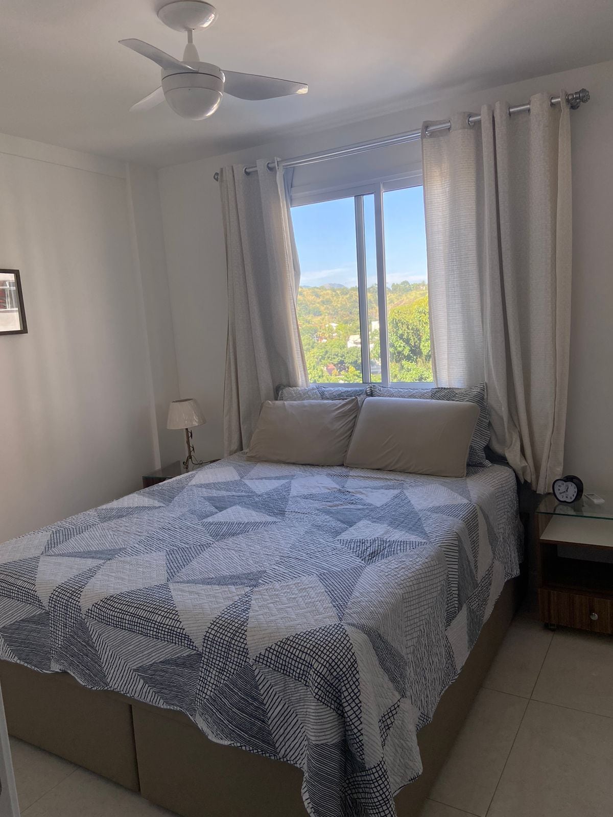 A cozy bedroom features a spacious bed covered with a geometric patterned blanket. Two neutral decorative pillows rest against the headboard. A small bedside table with a lamp sits beside the bed, while a window admits natural light, revealing a view of greenery outside.