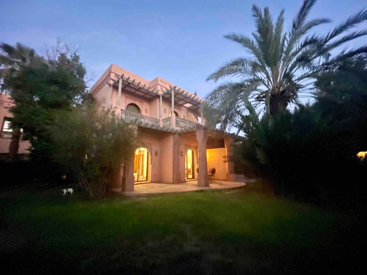 A charming villa is framed by lush greenery, showcasing a spacious patio area under a wooden pergola. The structure features multiple arched doorways and large windows, allowing for a seamless connection between indoor and outdoor spaces. Palm trees are visible in the background.