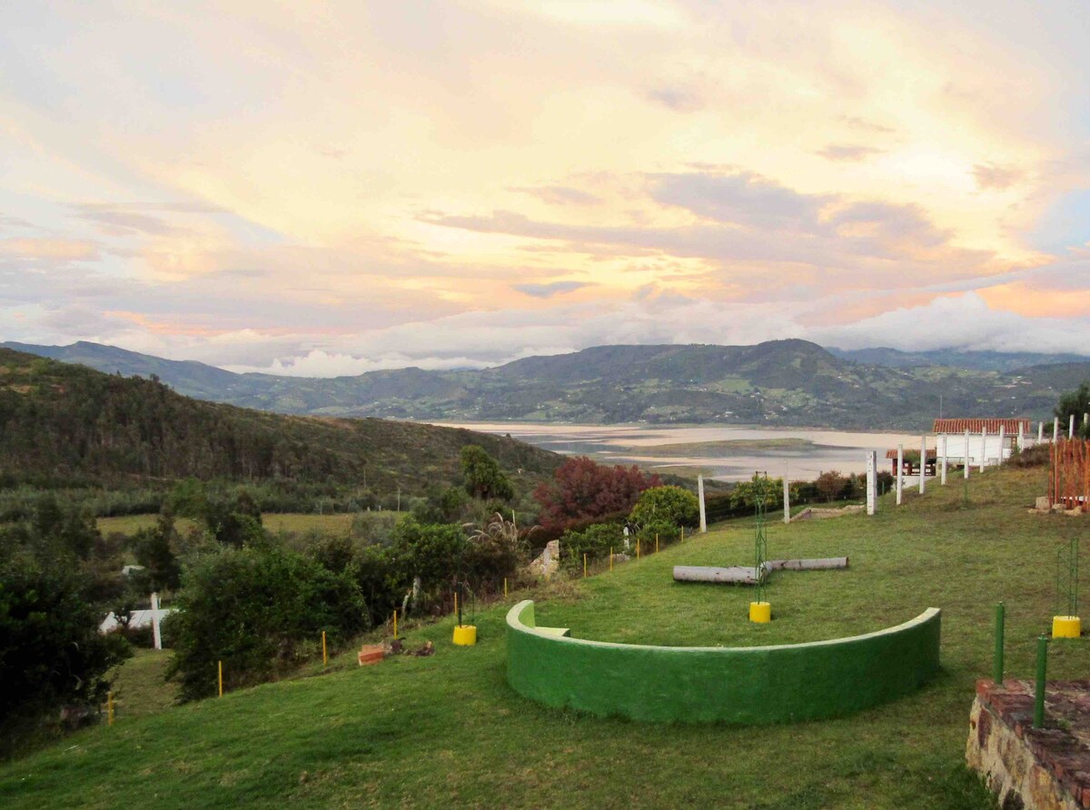 A spacious outdoor area features a curved green seating area overlooking a serene landscape. Natural greenery surrounds the scene, with rolling hills and a tranquil body of water visible in the distance. A colorful sky transitions from soft pastels to deeper hues at sunset.