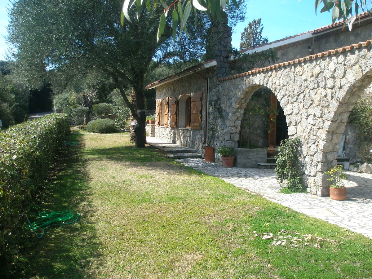 A charming stone façade is viewed against a backdrop of lush greenery. The entrance features arched doorways, and wooden shutters adorn the windows. Well-maintained grass and hedges frame the path, creating an inviting outdoor space surrounded by Mediterranean vegetation.