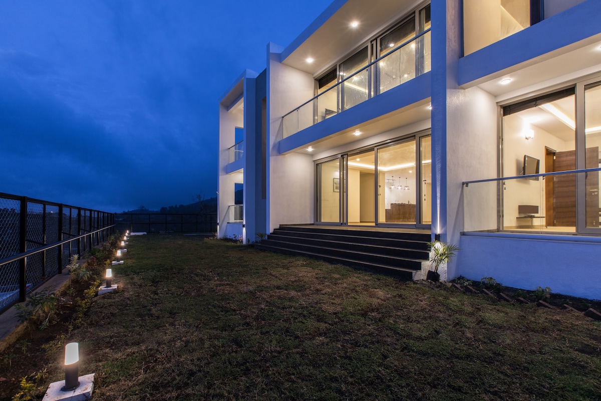The exterior of the guest house is illuminated at dusk, highlighting a modern architectural design. The entrance features multiple steps leading up to large glass doors, while pathway lights guide towards the entrance, surrounded by well-maintained grass and plants.