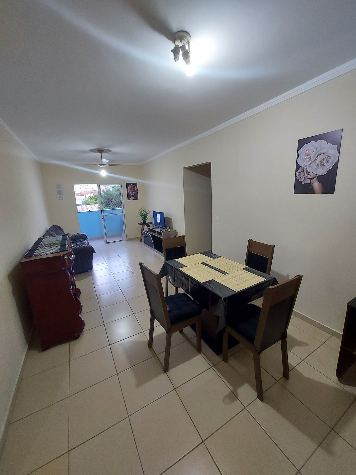 A well-lit living space features a dining table set for four, complemented by a wooden sideboard. A comfortable seating area is visible, with a television placed on a unit. Natural light enters through the balcony door, enhancing the inviting atmosphere.