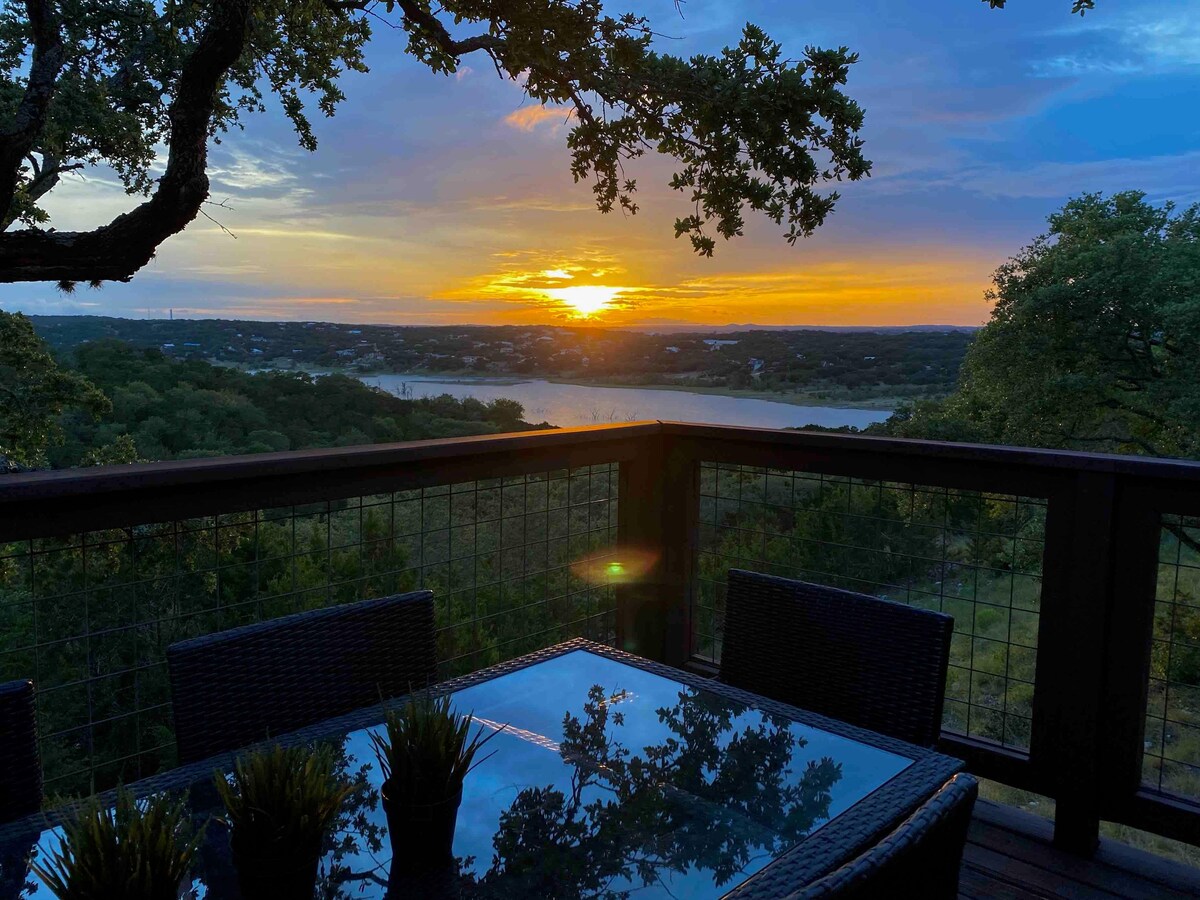 An outdoor dining area is depicted with a glass table and dark wicker chairs, framed by a backdrop of expansive hills. A colorful sunset casts warm tones across the sky, reflecting off the water of Canyon Lake in the distance.