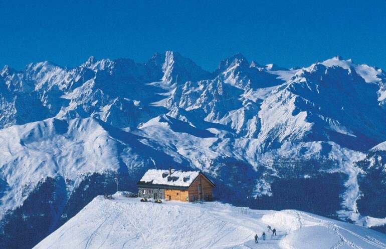 A snow-covered mountain scene features a wooden chalet situated on a hilltop, surrounded by expansive views of snow-capped peaks. The sky is clear and blue, complementing the serene winter landscape below.
