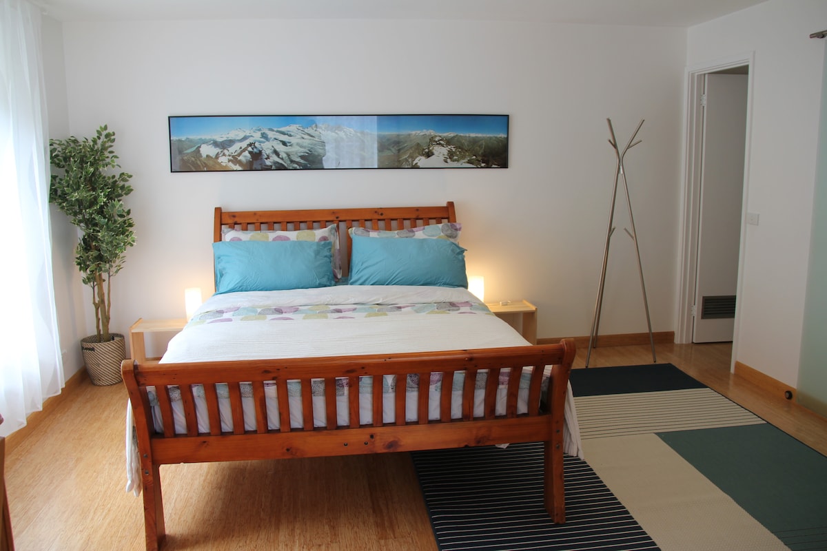 The bedroom features a wooden bed frame with a spacious mattress dressed in light-colored linens and decorative pillows. A panoramic landscape image of mountains hangs above the bed. Soft light is provided by bedside lamps, and natural light filters in through a sheer curtain.