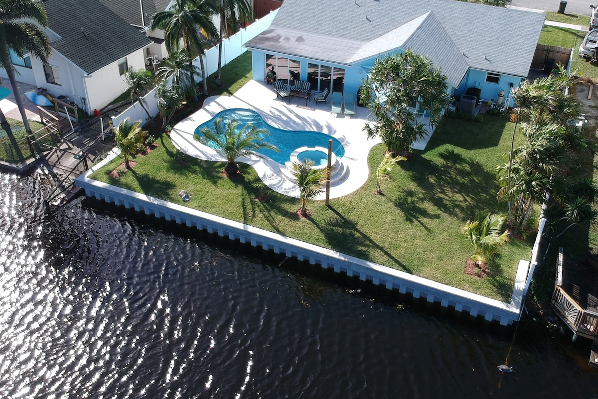 Aerial view illustrates the backyard oasis featuring a private pool surrounded by palm trees and manicured grass. The home is positioned near the water's edge, with a vibrant blue exterior visible in the background. The serene atmosphere is enhanced by the gentle waves of the river.