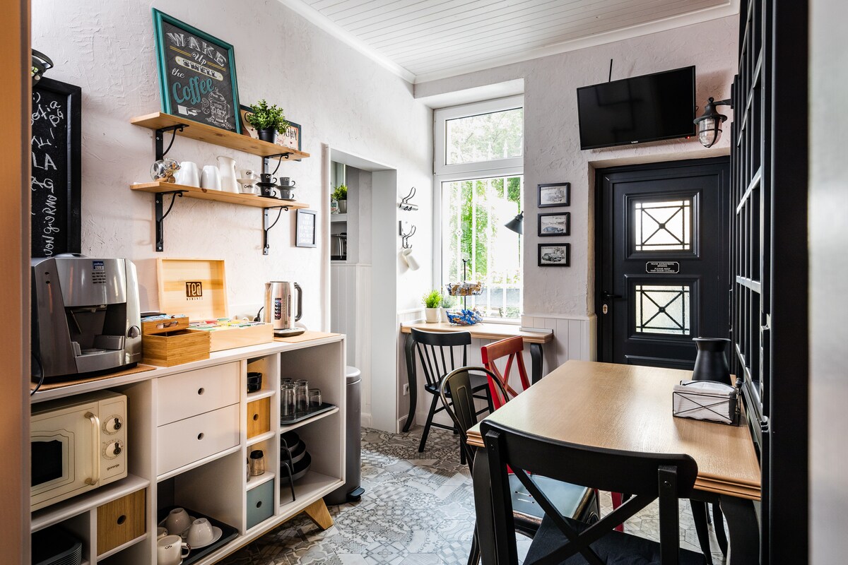 A bright and welcoming kitchen area features a small dining table and a coffee corner with a variety of appliances. Crafted wooden shelves display plants and decorative items. Natural light fills the space through a large window, and a door leads to the outside.
