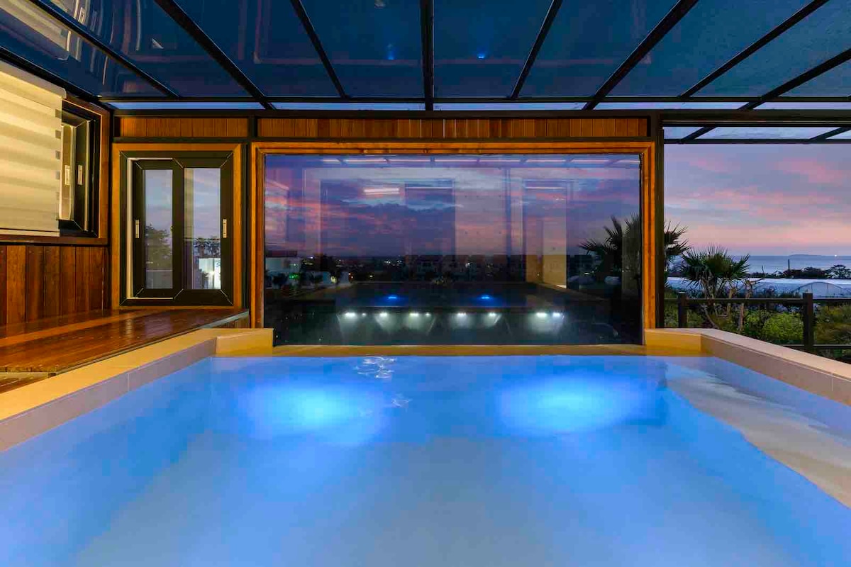 An indoor swimming pool features soft blue lighting, creating a serene ambiance. The pool is framed by large glass panels that offer a view of the sky and surrounding landscape, enhancing the sense of openness and relaxation.