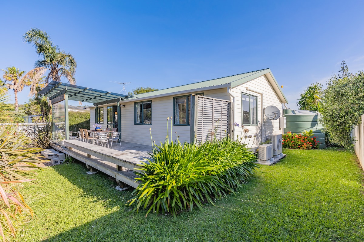 A single-story holiday home is set within a green landscape, featuring a spacious deck with outdoor seating. Lush plants frame the house, while large windows invite natural light. The surrounding lawn offers ample space for outdoor activities and is complemented by tropical palm trees.