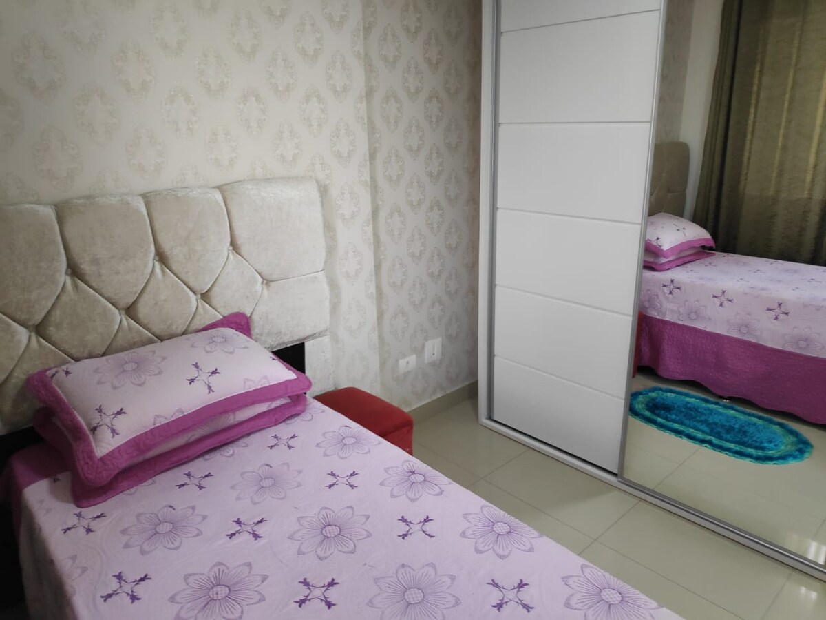 A tidy bedroom features a single bed dressed in pastel purple linens, accented with floral designs. A mirrored wardrobe reflects the space, while a plush area rug adds comfort. Natural light softly illuminates the room, enhancing its clean and organized appearance.