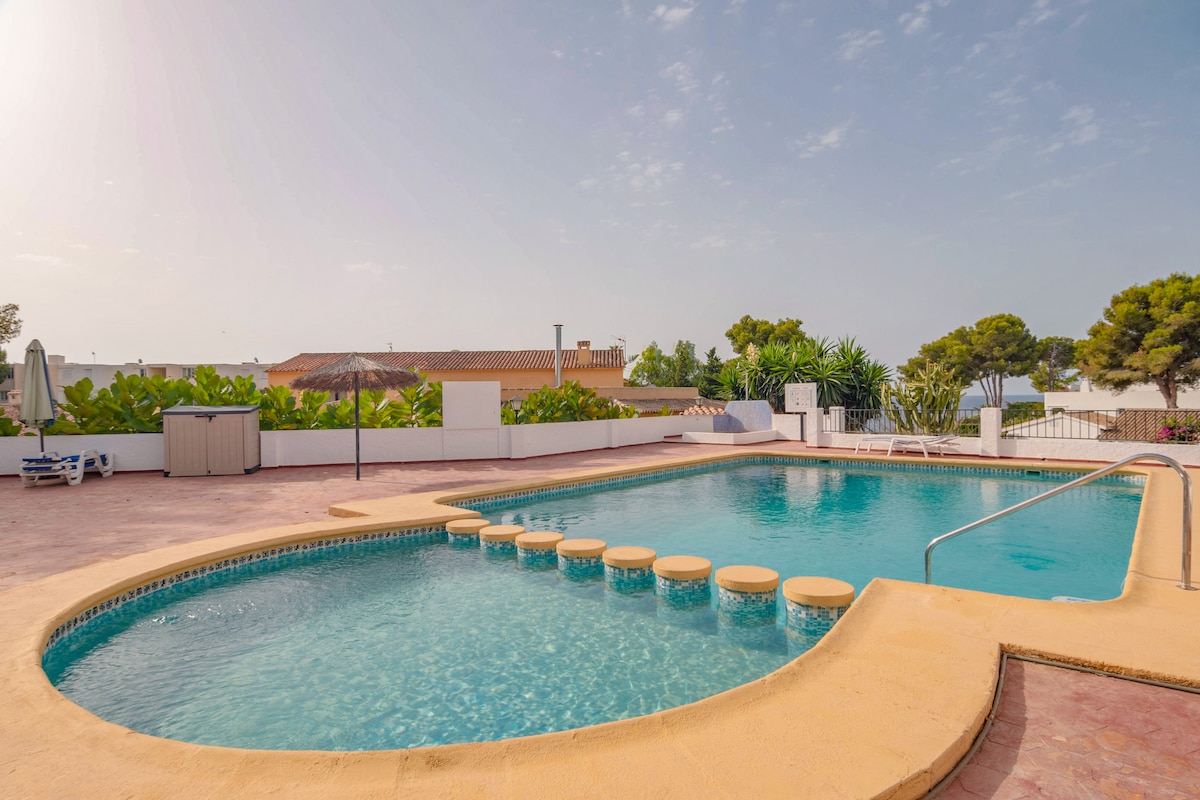 A communal swimming pool is featured, surrounded by well-maintained greenery. The pool's curved shape includes a series of sun loungers along one side. Clear blue water reflects the sky, while a shaded area provides cover from the sun, creating a relaxing outdoor environment.