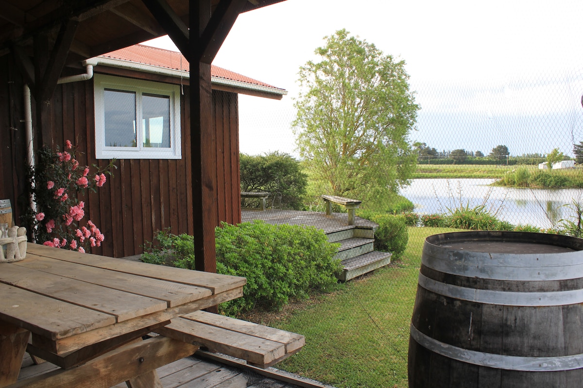 A wooden dining table is situated under a covered porch, with a view of a landscaped garden leading down to a peaceful body of water. Blooming flowers and a rustic barrel add charm to the setting, enhancing the connection to nature.