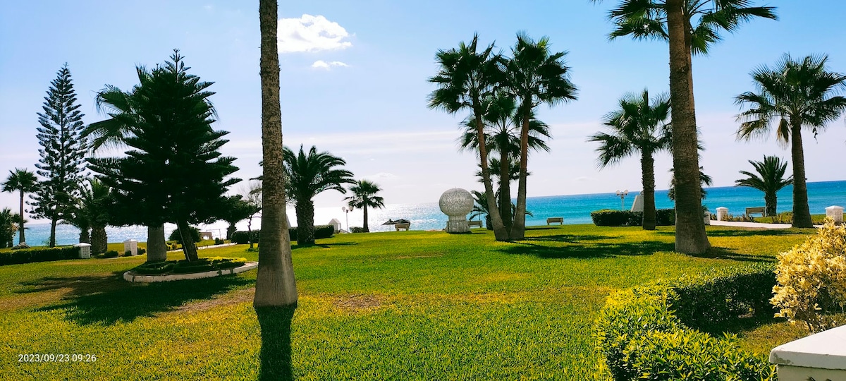 A landscaped garden features lush green grass and palm trees, providing a serene environment. The ocean is visible in the background, and several seating areas are positioned throughout the space, inviting relaxation by the water.