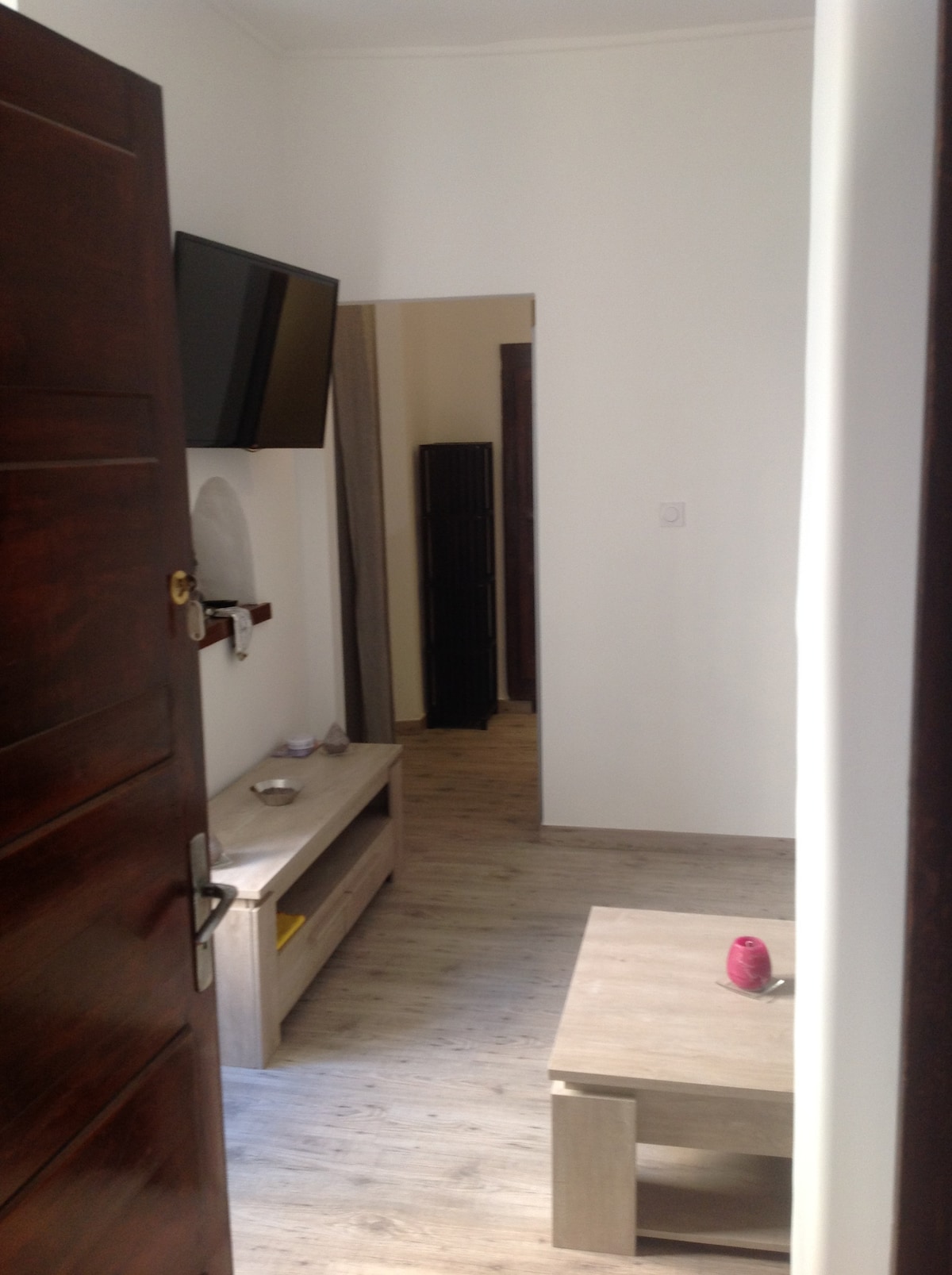 A clean and bright entrance reveals a compact living area featuring a mounted television above a low cabinet. A wooden table sits centrally, and soft lighting highlights the neutral-colored walls and wooden floor. A doorway leads to additional living spaces.