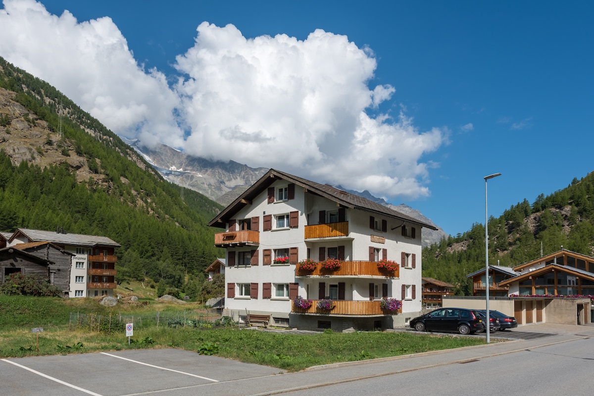 A three-story chalet-style building is set against a backdrop of mountains and blue skies, with fluffy white clouds. The structure features several balconies adorned with colorful flower boxes. A parking area is visible in the foreground, and additional rustic buildings are seen nearby.