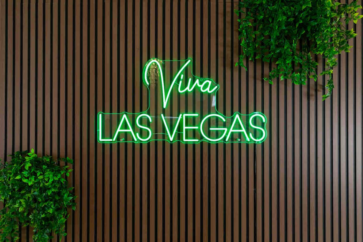 A vibrant neon sign reading 'Viva Las Vegas' glows in bright green against a textured wooden wall. The wall is complemented by a small, lush plant in the corner, adding a touch of greenery to the lively atmosphere.