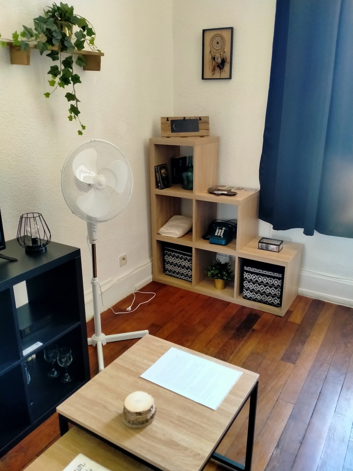 A well-organized living area features a small wooden table and a stand fan for comfort. Shelves display decorative items and neatly folded towels, while a dark cabinet holds glassware. Light streams in through a blue curtain, enhancing the natural wood flooring.