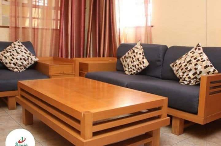 A comfortable living area features two blue sofas adorned with patterned cushions, arranged around a wooden coffee table. Natural light enters through large windows, complemented by soft red curtains. Warm tones of the furnishings create a welcoming environment.