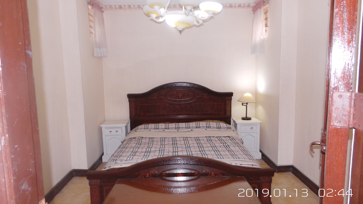 An inviting bedroom features a spacious bed with a plaid blanket, flanked by two nightstands. A warm lamp provides soft lighting, and light filters in through sheer curtains on the window, enhancing the cozy atmosphere of the room.