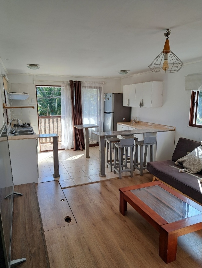 A bright living area features a cozy futon, a glass coffee table, and modern stools around a small dining counter. Natural light fills the space through large windows, which open to a balcony. The kitchen area is equipped with cabinetry and appliances, displaying a clean and functional design.
