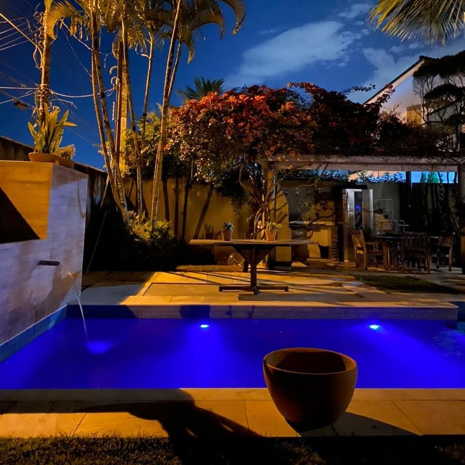 A serene outdoor space is highlighted by a well-lit swimming pool, displaying calming blue hues. Surrounding palm trees and blooming plants add natural beauty. A dining table and chairs are positioned nearby, creating an inviting area for relaxation and gathering.
