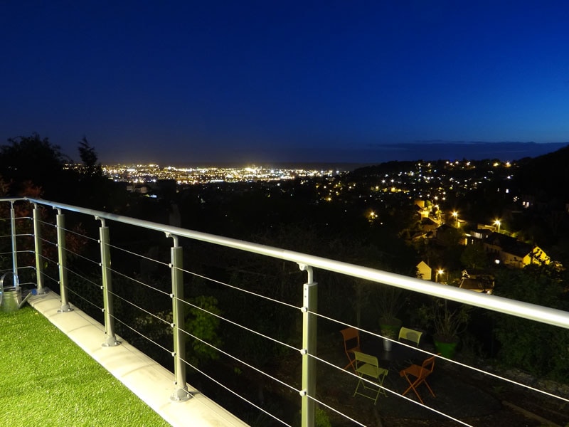 A terrace railing frames a breathtaking panoramic view of Rouen at night, with city lights twinkling against a deep blue sky. Seating is arranged nearby on the lush, green grass, inviting moments of relaxation while enjoying the serene atmosphere.