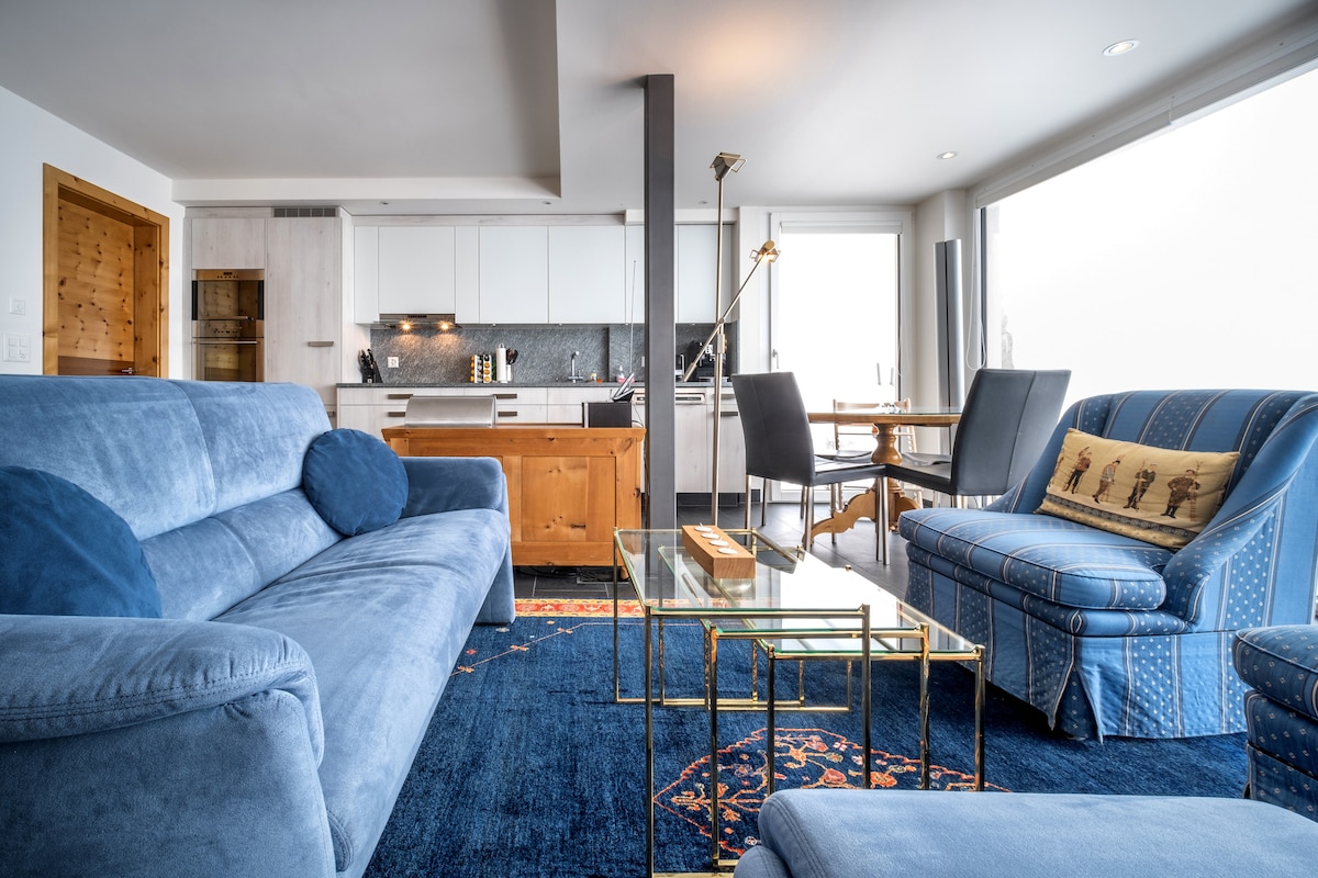 A spacious living area features plush blue sofas arranged around a glass coffee table, with a decorative carpet underneath. The modern kitchen is visible in the background, complemented by a dining table positioned near large windows that allow natural light to illuminate the space.