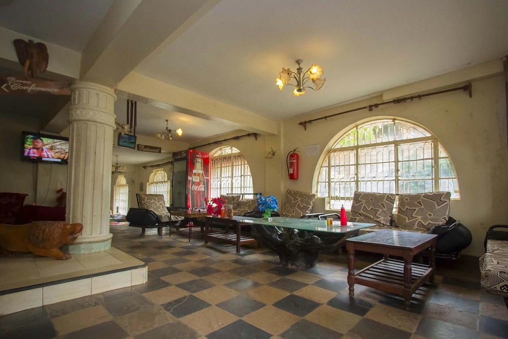 A spacious lounge area is highlighted by large arched windows, allowing natural light to enter. Comfortable seating is arranged around a central coffee table. Decorative elements, such as a TV on the wall and potted flowers, add character to the room.