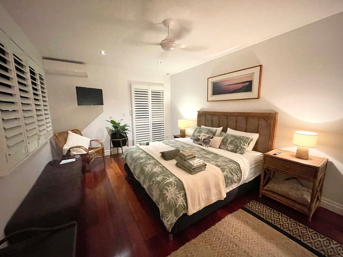 A spacious bedroom features a comfortable bed with decorative pillows and soft linens. Natural light enters through shuttered windows, complementing the warm wood tones. A wicker chair, small side table, and a flat-screen TV are present. Subtle artwork hangs above the bed, adding a touch of character.