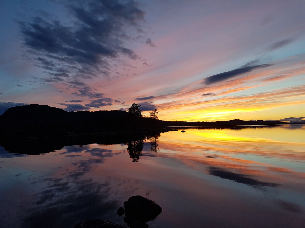 A serene landscape is captured during sunset, with vibrant colors reflecting off the calm water. Soft hues of orange, pink, and purple fill the sky, while silhouettes of trees and distant hills are visible along the horizon, creating a tranquil atmosphere.