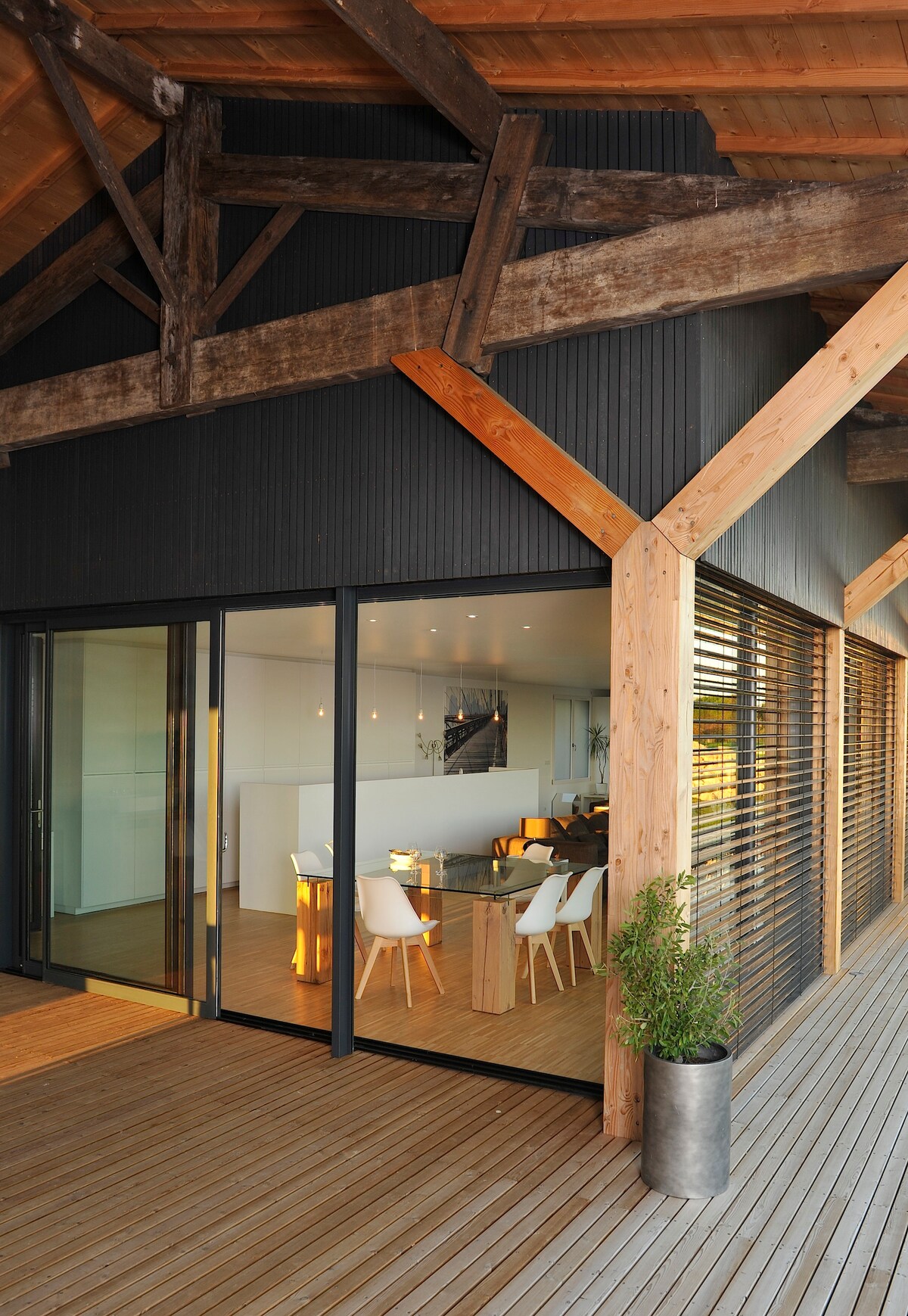 An expansive outdoor deck features wooden flooring, seamlessly connecting to glass doors. Interior dining space is visible, showcasing modern furnishings including white chairs and wooden tables under a wood-beamed ceiling. Natural light enhances the atmosphere, supported by a planted container on the deck.
